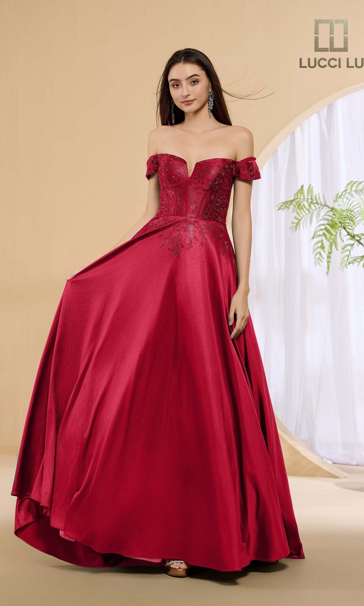 A woman stands before a neutral background wearing the Lucci Lu 1483, a wine-colored long prom dress featuring an off-the-shoulder corset bodice and flowing floor-length stretch satin skirt.